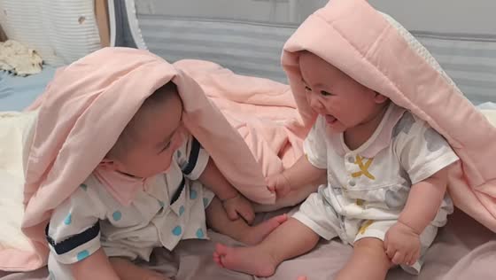 Twin babies laugh together under blanket on bed in Hubei, China