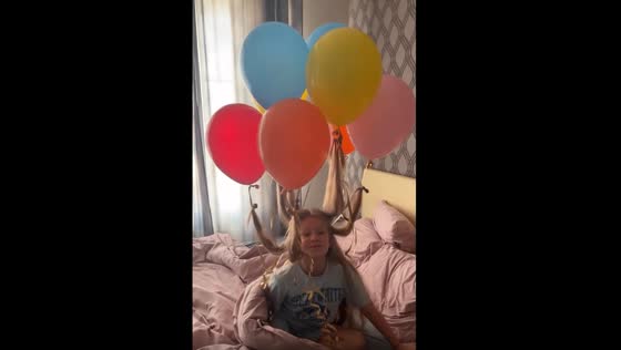 A girl with floating balloons tied to her hair is waiting for her brother's birthday!